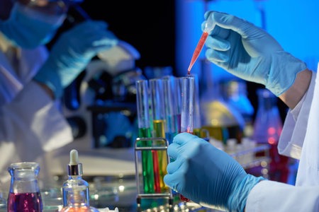 Talented researcher wearing rubber gloves standing at laboratory bench and pouring red liquid into...