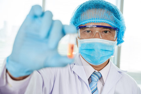 Asian middle-aged microbiologist wearing white coat and safety goggles holding brand-new vaccine in...