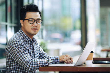 Portrait of smiling Asian man working on laptop in cafeの写真素材