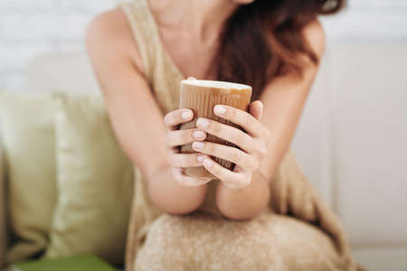Close-up of unrecognizable tanned woman sitting on sofa and holding beautiful brown ceramic mug while drinking teaの写真素材