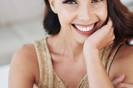 Close-up of joyful excited emotional young woman with white teeth and sensual lips leaning on hand and smiling at cameraの写真素材