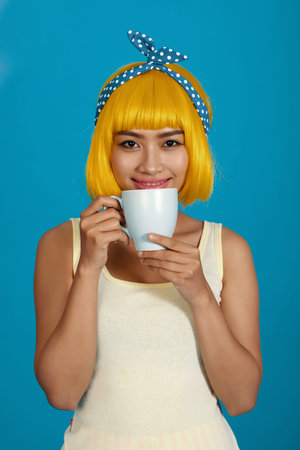 Cheeful young woman in yellow wig drinking coffee