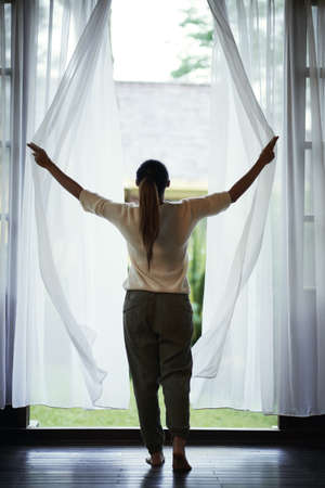 Young woman standing by window and opening curtainsの写真素材
