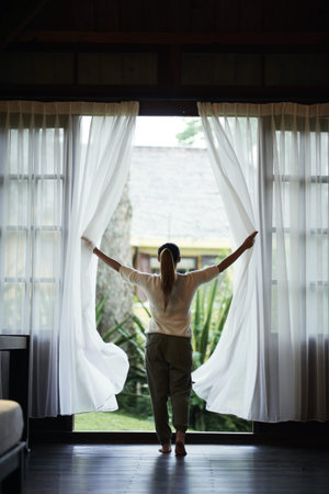 Rear view of woman opening curtains to see a beautiful viewの写真素材