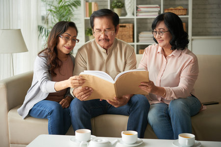 Three senior friends sitting on sofa and looking at family album and drinking tea at homeの写真素材