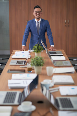 Waist-up portrait of confident Asian entrepreneur wearing elegant suit leaning on wooden table and posing for photography with wide smile, interior of modern boardroom on backgroundの写真素材