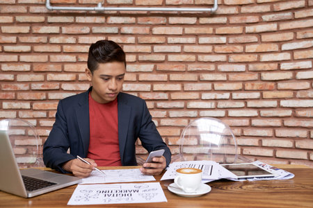 Waist-up portrait of handsome Vietnamese manager sitting at wooden desk and finishing ambitious...