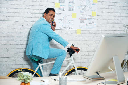 Stylish businessman in light blue suit sitting on bicycle in office and talking on cell phoneの写真素材
