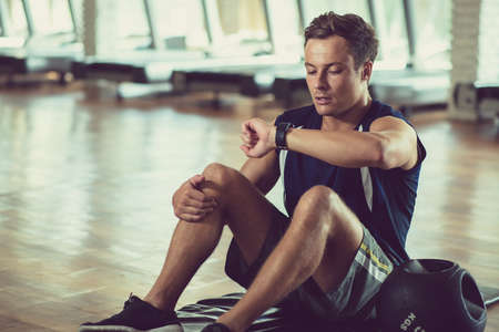 Full length portrait of handsome sportsman sitting on mat and checking heart rate with help of fitness tracker after completion of intensive trainingの写真素材