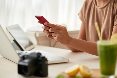 Close-up shot of unrecognizable woman sitting at kitchen table and texting with friend on smartphone while taking short break from work on food blogの写真素材
