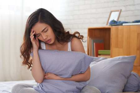 Portrait shot of beautiful Asian woman sitting on bed and holding pillow in hand while suffering...