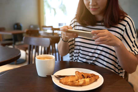 Smiling woman wearing striped T-shirt sitting at cafe table and taking picture of appetizing croissant and fragrant coffee on smartphoneの写真素材