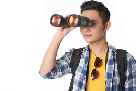 Portrait shot of handsome Asian tourist with backpack looking through binoculars while standing against white backgroundの写真素材