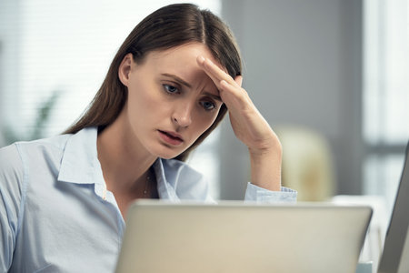 Young puzzled woman leaning on hand while using computer at workplace having problems with...