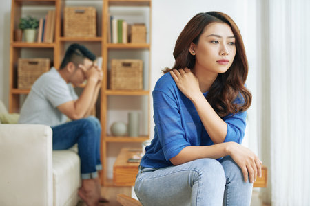 Sad Asian woman sitting alone with man on background being in conflict at home