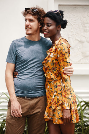 Black woman and Caucasian man in summer outfits standing and embracing outside while looking away dreamilyの写真素材