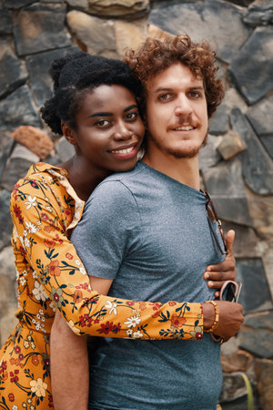 Charming African-American woman embracing Caucasian bearded man from back both looking at camera with smileの写真素材