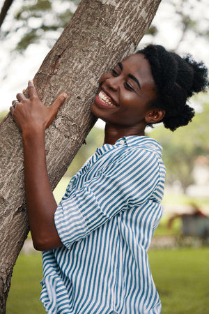Side view of laughing African-American woman embracing tree trunk in park enjoying nature
