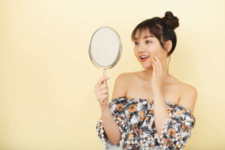 Pretty Asian girl looking at the mirror isolated on beige backgroundの写真素材