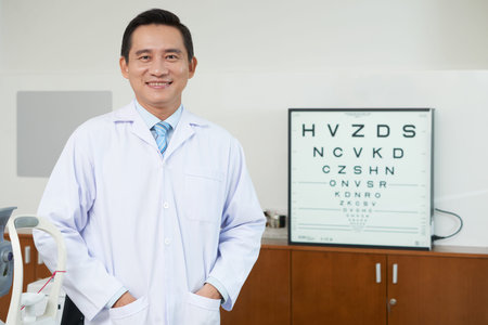 Cheerful Asian man in uniform of medic standing in ophthalmology cabinet with eye test letters on...