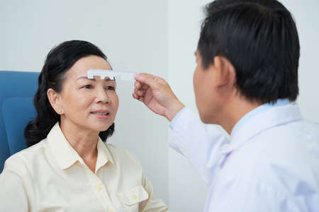 Professional oculist using special ruler and measuring eyes of mature Asian woman in hospital cabinetの写真素材