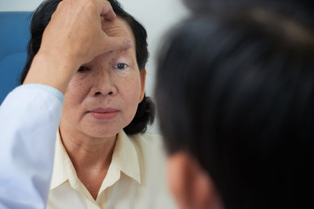Crop doctor flashing with light into eye of mature Asian woman checking pupil reaction while testing...