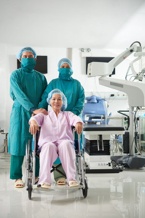 Mature Asian woman sitting in wheel-chair with professional surgery doctors near looking at camera