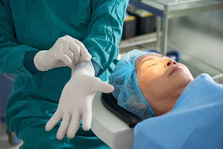 Faceless surgeon putting on sterile gloves with mature woman lying on operating table before eye...