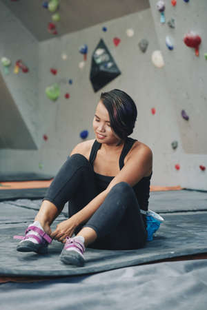 Asian brunette female in sportswear and insurance equipment on belt sitting on mat putting on trainers in climbing clubの写真素材