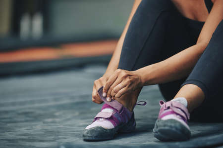 Crop sportive female putting on sneakers sitting on mat in gym on blur backgroundの写真素材