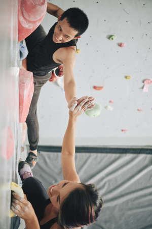 From above shot of happy Asian man and woman giving high five while hanging on clambering wall in gymの写真素材