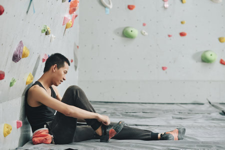Side view of Asian young man in black sportswear tying shoelace of footwear sitting on floor under climbing wallの写真素材