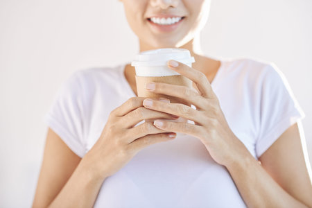 Crop smiling female in white t-shirt holding takeaway coffee in hands on light background