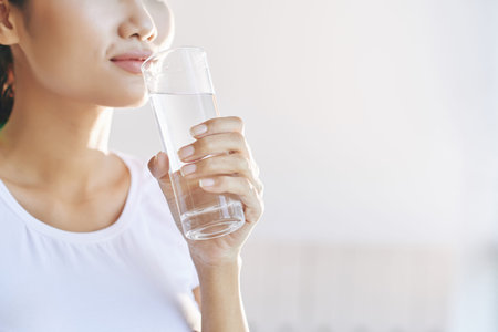 Crop beautiful female in white t-shirt drinking water holding glass in hand on light blur background