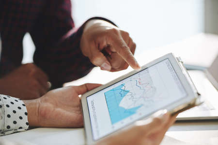 Close-up shot of crop man pointing at diagram on tablet screen while having discussion with colleague at workplaceの写真素材