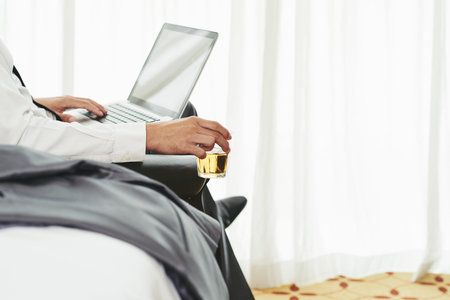 Businesman drinking whiskey and checking e-mails on laptopの写真素材