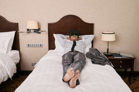 Businessman relaxing on bed in hotel room and enjoying virtual realityの写真素材