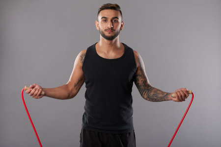 Handsome tattooed man in black sportswear looking seriously at camera while jumping rope on gray...