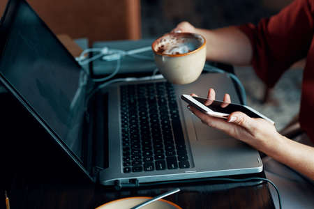 Close-up of woman connecting mobile phone to the laptop and drinking coffeeの写真素材
