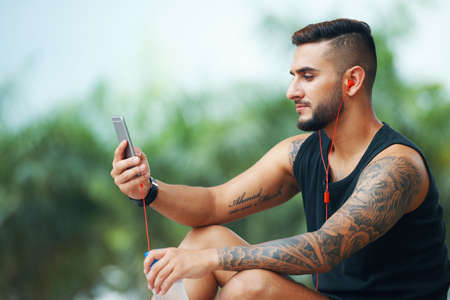 Handsome adult tattooed man in headphones using smartphone while sitting with bottle of water outdoorsの写真素材