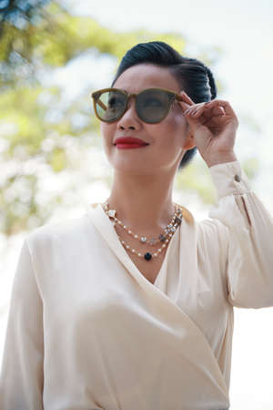 Beautiful cheerful Asian female in delicate blouse and necklace touching trendy sunglasses and looking away standing outdoors on sunny summer day on blurred backgroundの写真素材