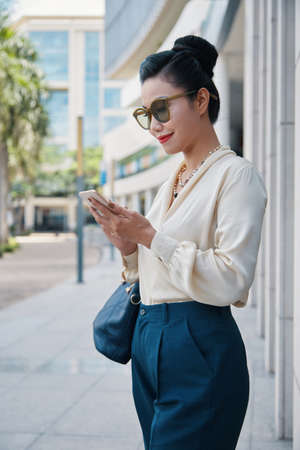 Side view of gorgeous Asian female in elegant clothes and sunglasses holding handbag using mobile phone and smiling standing on pavement at building in summer on blurred background.の写真素材