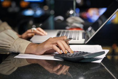 Crop side view of female hands in light blouse sitting at dark desk with laptop and papers and using...