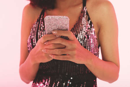 Crop view of woman in black top with silver sequins holding in hands mobile phone covered with silver glitter over pink studio backgroundの写真素材
