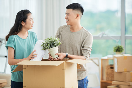 Beautiful cheerful ethnic woman and man in casual clothes putting plant into large open cardboard...