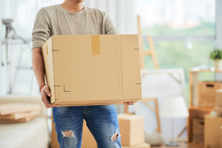 Crop view of strong male in casual clothes carrying large cardboard box on moving day with cartons...