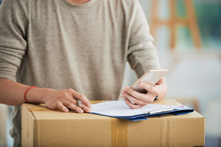 Crop view of man in casual clothes standing with pen at delivered packed cardboard box with clipboard and working with mobile phone in hand on blurred background .の写真素材