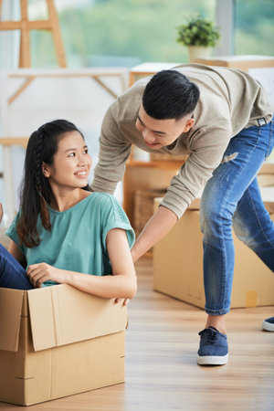 Beautiful young Asian woman in casual clothes sitting in large cardboard box and looking happily at...