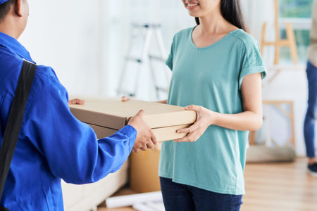 Crop view of happy female in casual clothes taking boxes from pizza delivery person in blue coat during break on moving day with cartons on blurred background .の写真素材