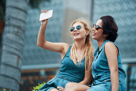 Attractive young women in sunglasses sitting and taking selfie with smartphone on streetの写真素材